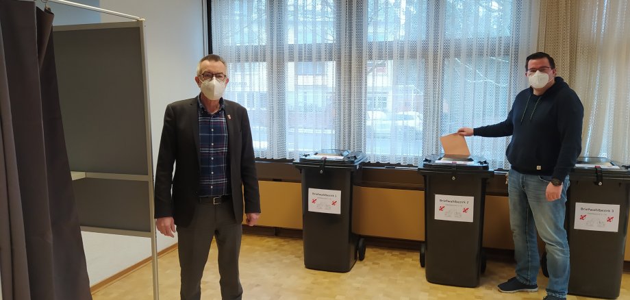Jirasek und Sander im Wahlbüro vor Wahlurnen und Wahlkabine.