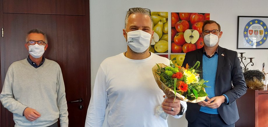 Drago Knezevic mit Blumenstrauß neben Bürgermeister Seitz und dem Ersten Beigeordneten Franz Jirasek. Alle tragen Masken.