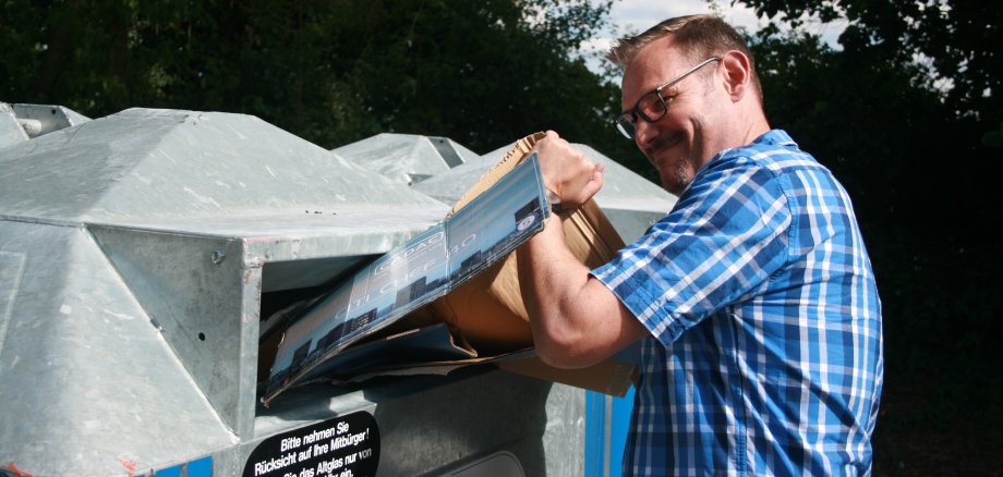Bürgermeister Seitz versucht einen Karton in einen vollen Papiercontainer zu pressen.