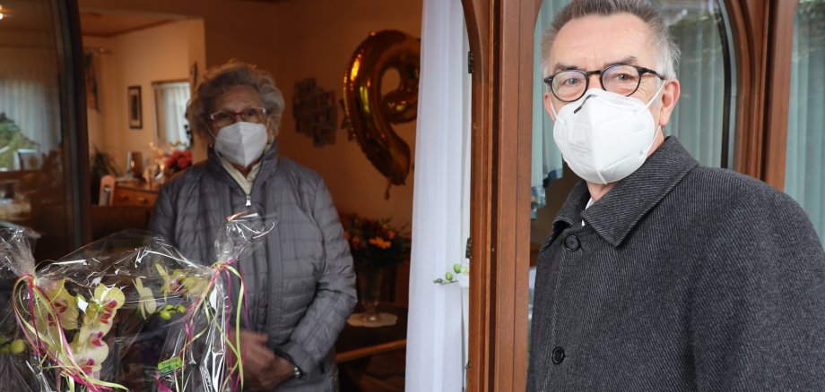 Anita Schäfer steht mit Maske und Jacke im Haus, Franz Jirasek mit Maske und Blumen draußen vor dem Fenster.