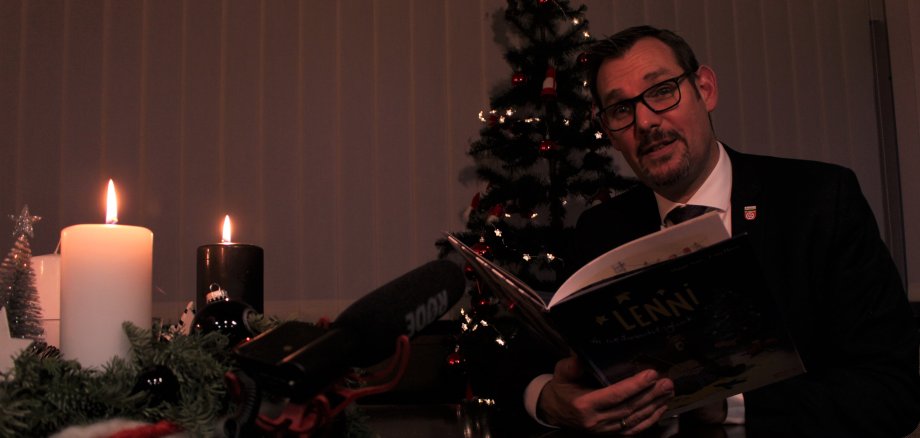 Bürgermeister Seitz mit Bilderbuch vor einem Tannenbaum in seinem Büro.