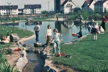 Kinder spielen im Weiher. Ringsherum freie Fläche.