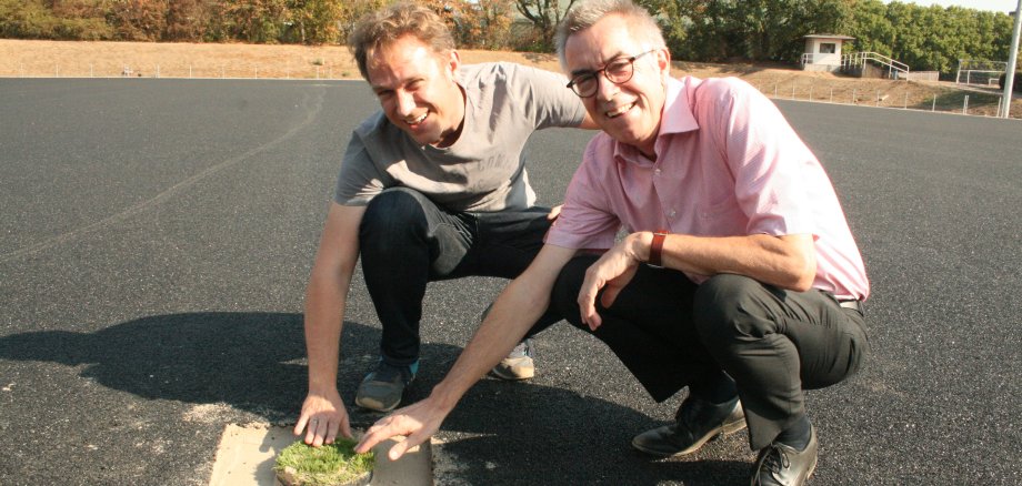 Franz Jirasek und Martin Mohr vom Bauamt streicheln erstes Grün auf dem Kunstrasenplatz.